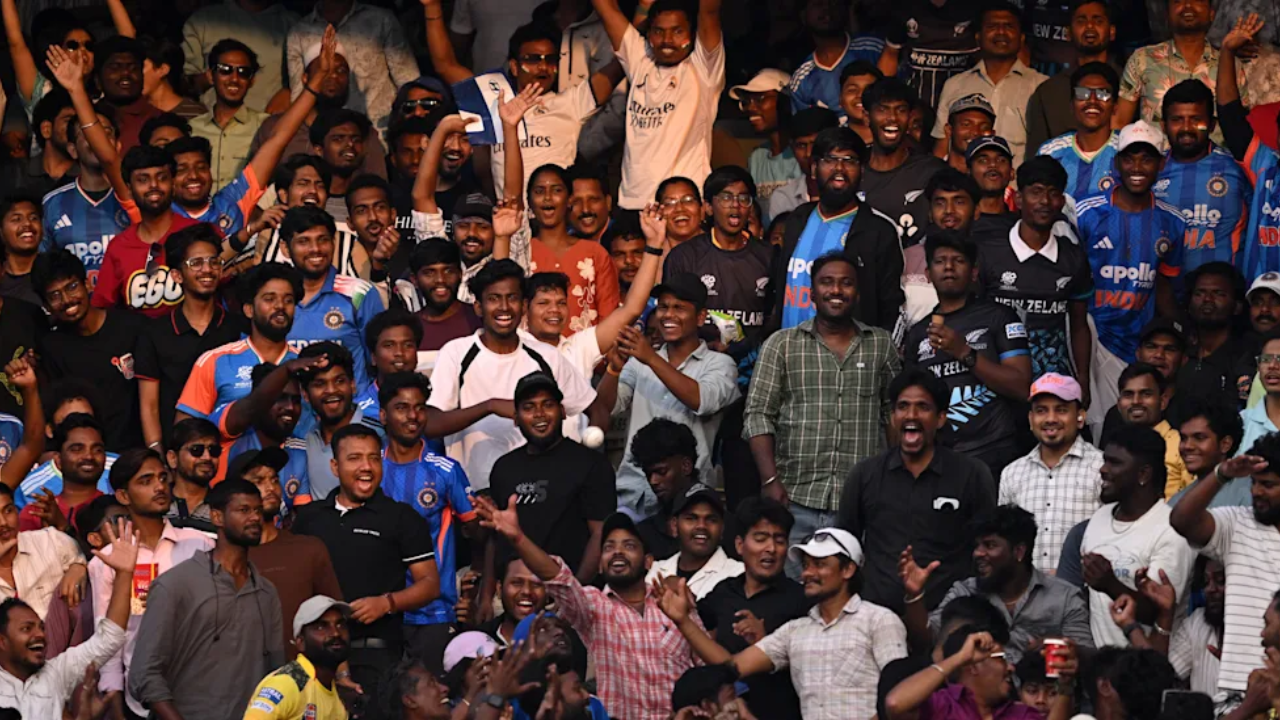 Cricket fans wearing India and New Zealand jerseys cheering enthusiastically in a packed stadium during an international match.