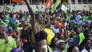 Jamaican track and field fans cheering in a packed stadium, waving flags and holding up scorecards during an athletics competition.