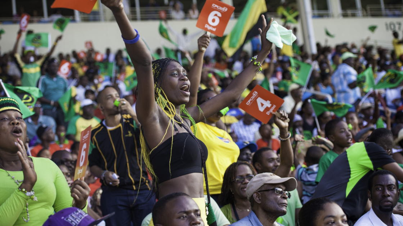 Jamaican track and field fans cheering in a packed stadium, waving flags and holding up scorecards during an athletics competition.