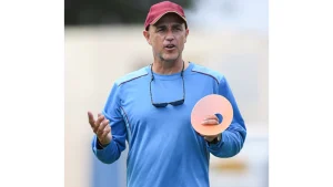 Cricket coach Richard Pybus conducting a training session, wearing a blue top and red cap while holding a practice cone on the field.