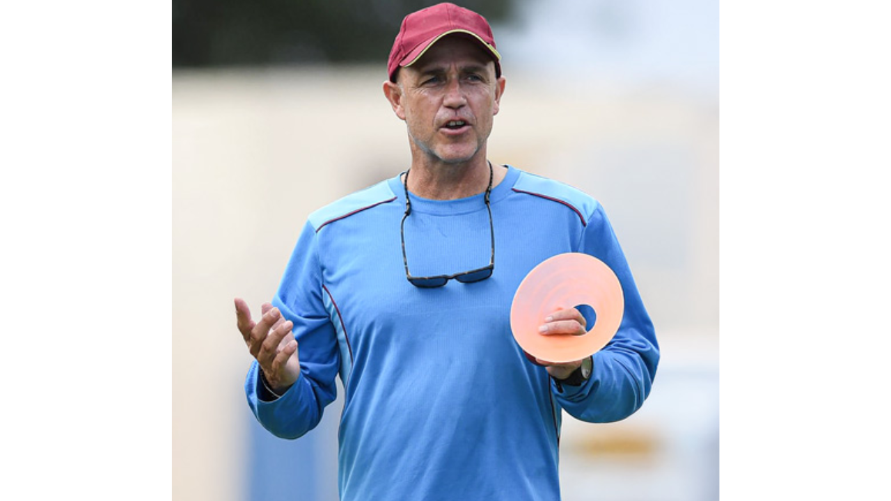 Cricket coach Richard Pybus conducting a training session, wearing a blue top and red cap while holding a practice cone on the field.