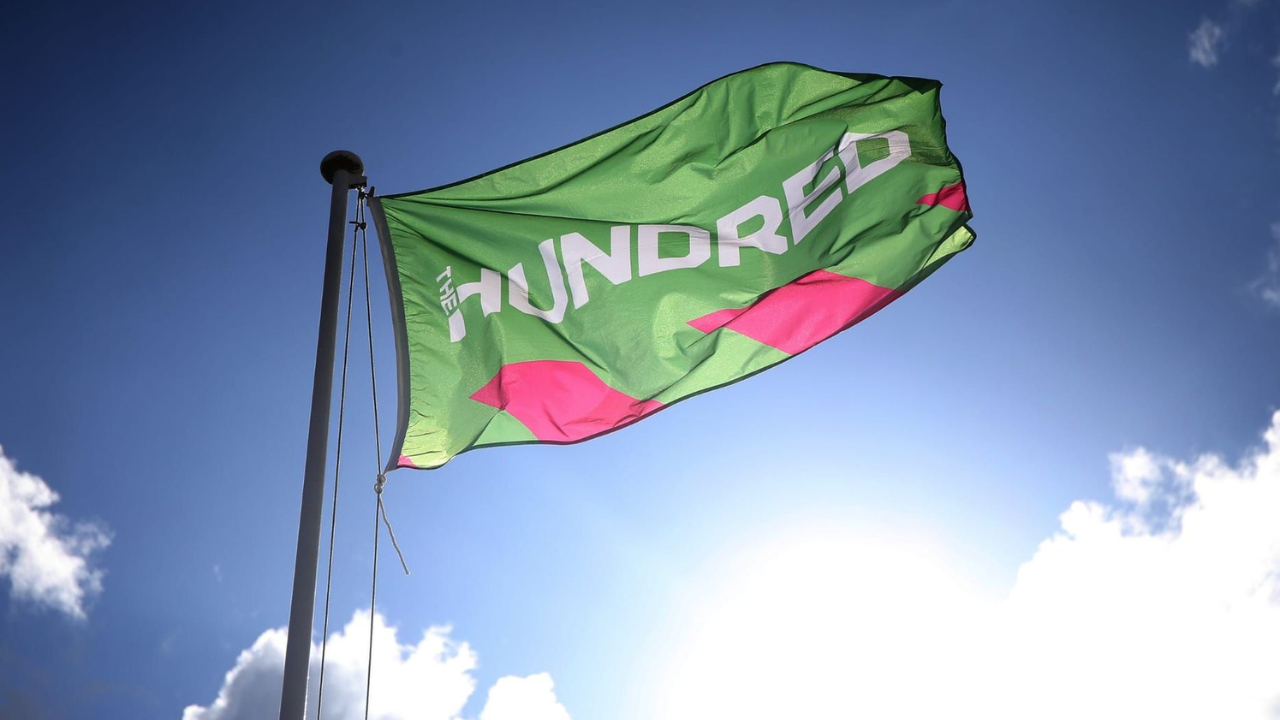 The Hundred cricket competition flag flying on a pole against a blue sky at a stadium.