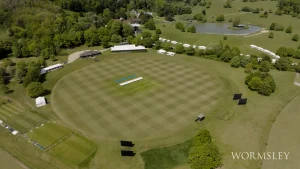 Aerial view of the Wormsley cricket ground in England surrounded by lush greenery, practice nets, and white marquees