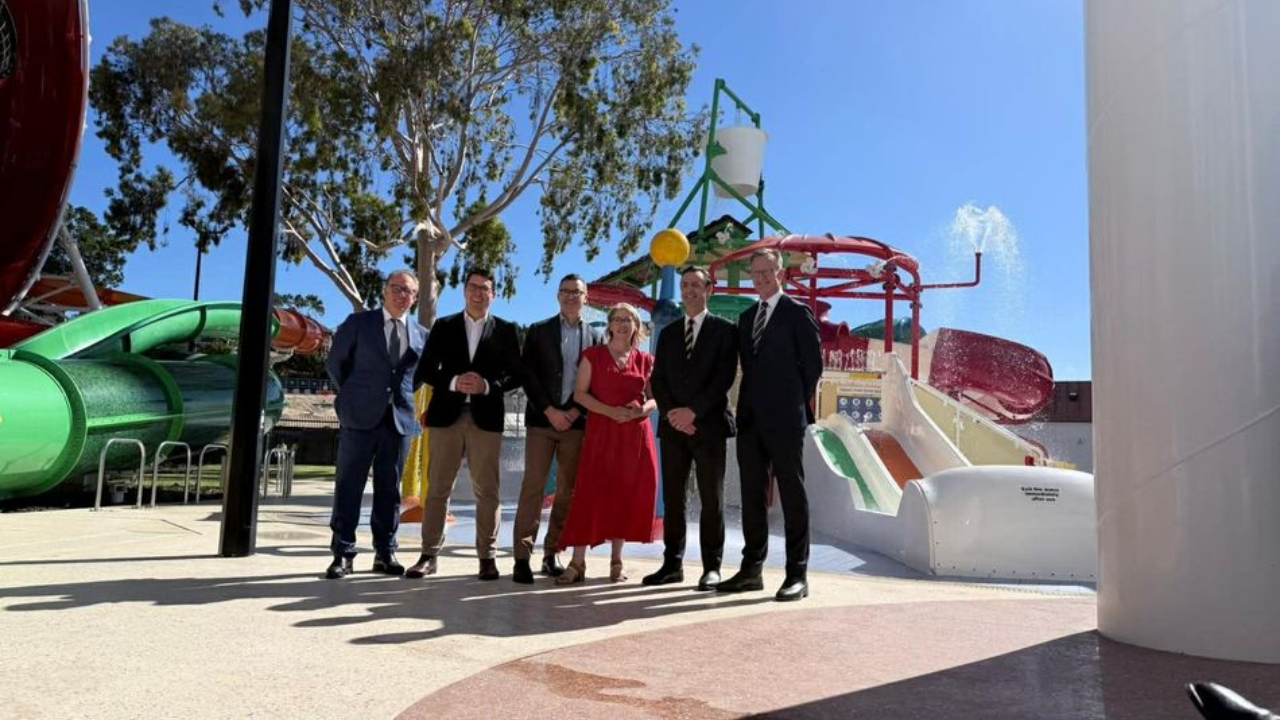 Government officials and executives pose during a site visit at a newly developed city water park facility with slides and splash structures in the background.