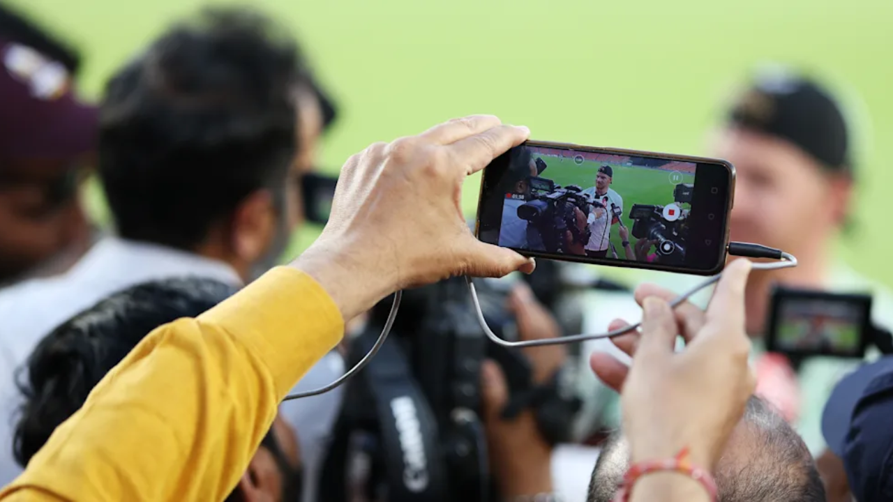 Journalists and cameramen recording a cricket player interview during a media interaction, with a smartphone capturing the press conference on a cricket field.
