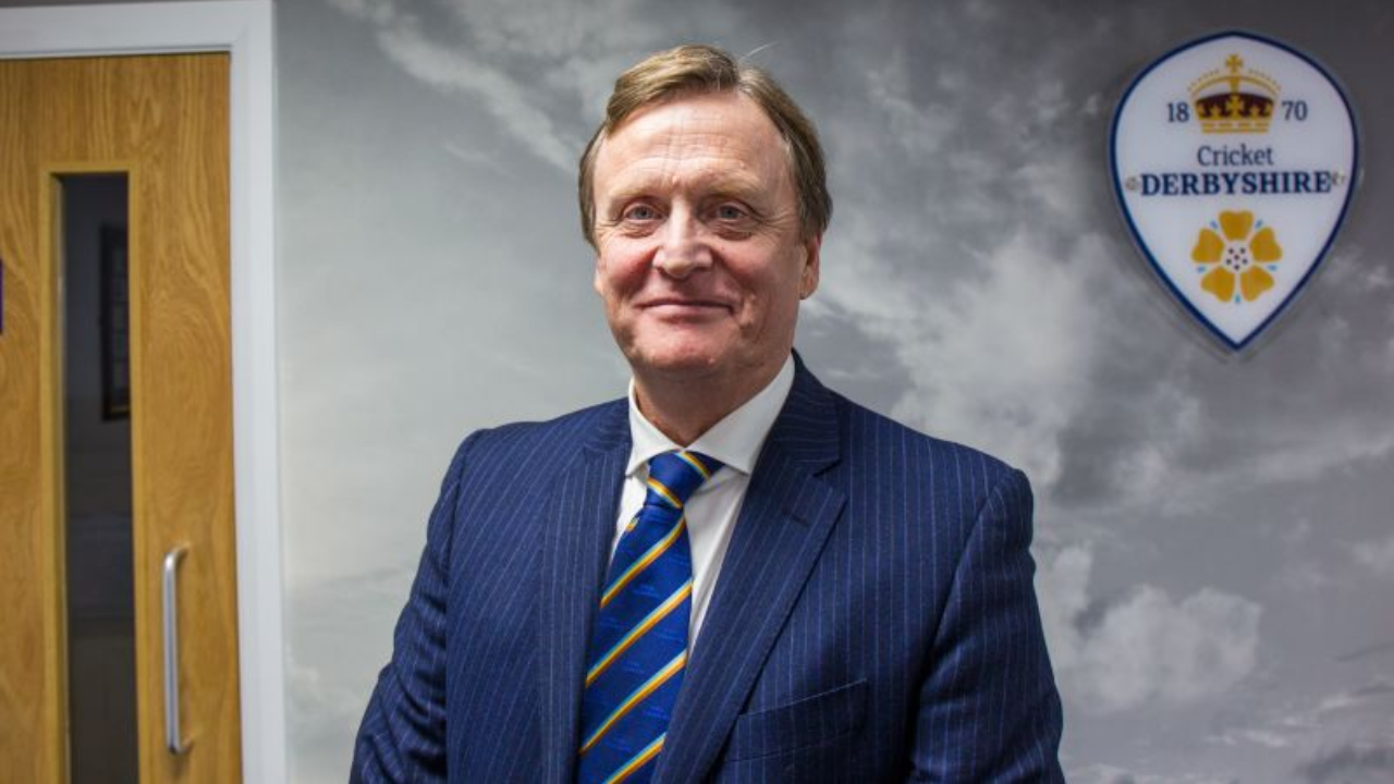 Ian McFarlane, Chair of Derbyshire County Cricket Club, posing in formal attire with club logo in the background