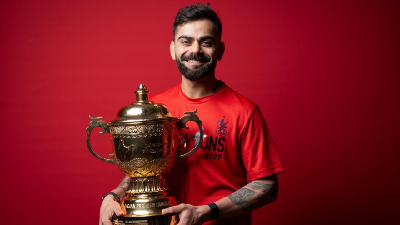 Virat Kohli holding the IPL trophy while wearing a Royal Challengers Bengaluru champions shirt against a red background.