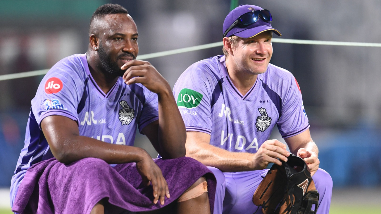 Andre Russell and Shane Watson sitting in the dugout as KKR coaches during an IPL match, wearing team jerseys and watching the game attentively