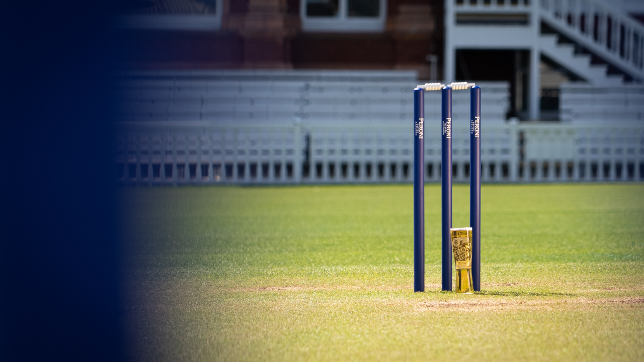Close-up of cricket stumps and bails on a grass pitch with pavilion seating blurred in the background.