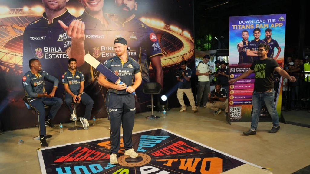 Gujarat Titans player Glenn Phillips participating in a fan batting activity on stage during a live promotional event with teammates and audience in background