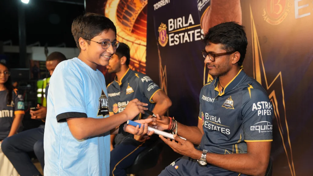 Sai Sudharsan signing an autograph for a young fan during a Gujarat Titans meet and greet event with team branding in the background