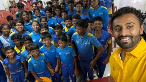Indian cricketer Shreyas Gopal taking a selfie with a large group of young cricket academy players wearing blue uniforms indoors.