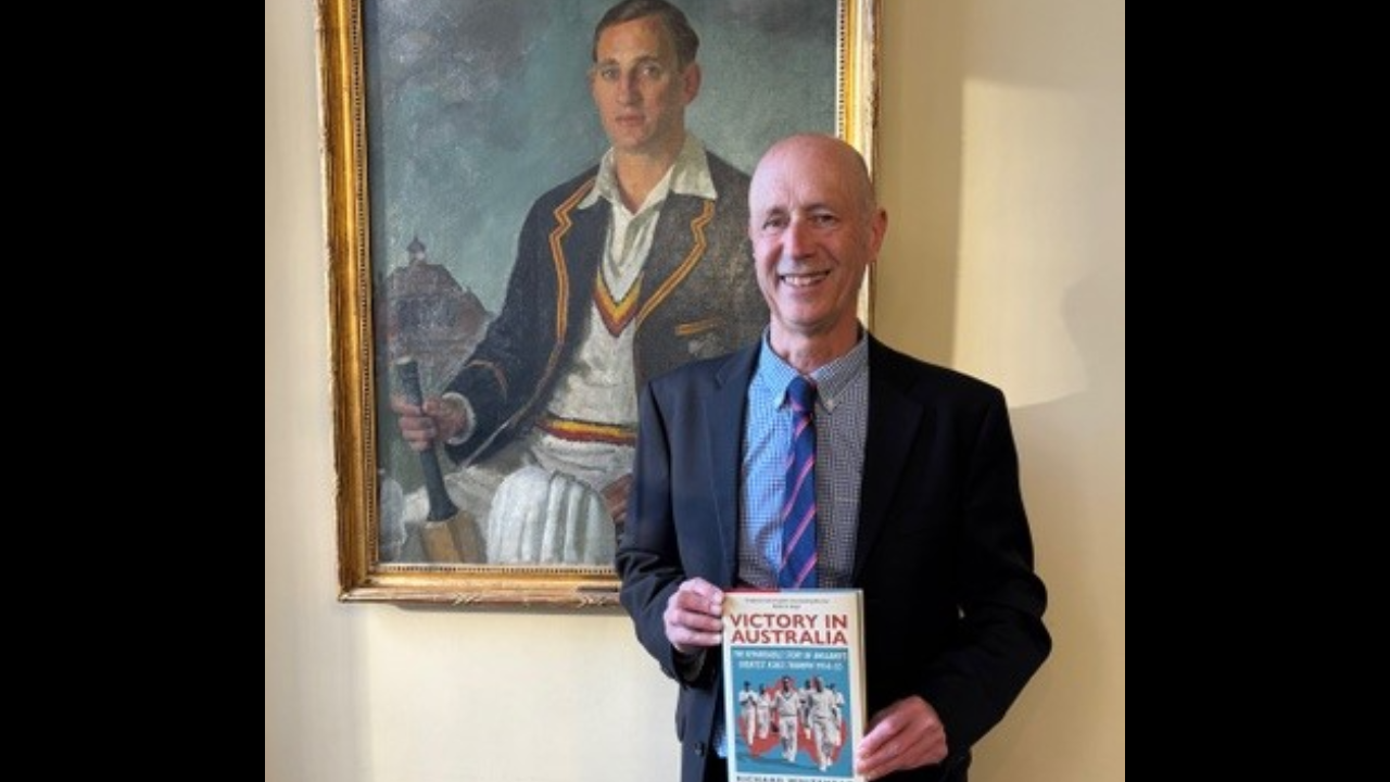 Author Richard Whitehead holding his book “Victory in Australia,” winner of the Cricket Society and MCC Book of the Year Award, standing beside a historic cricket portrait.