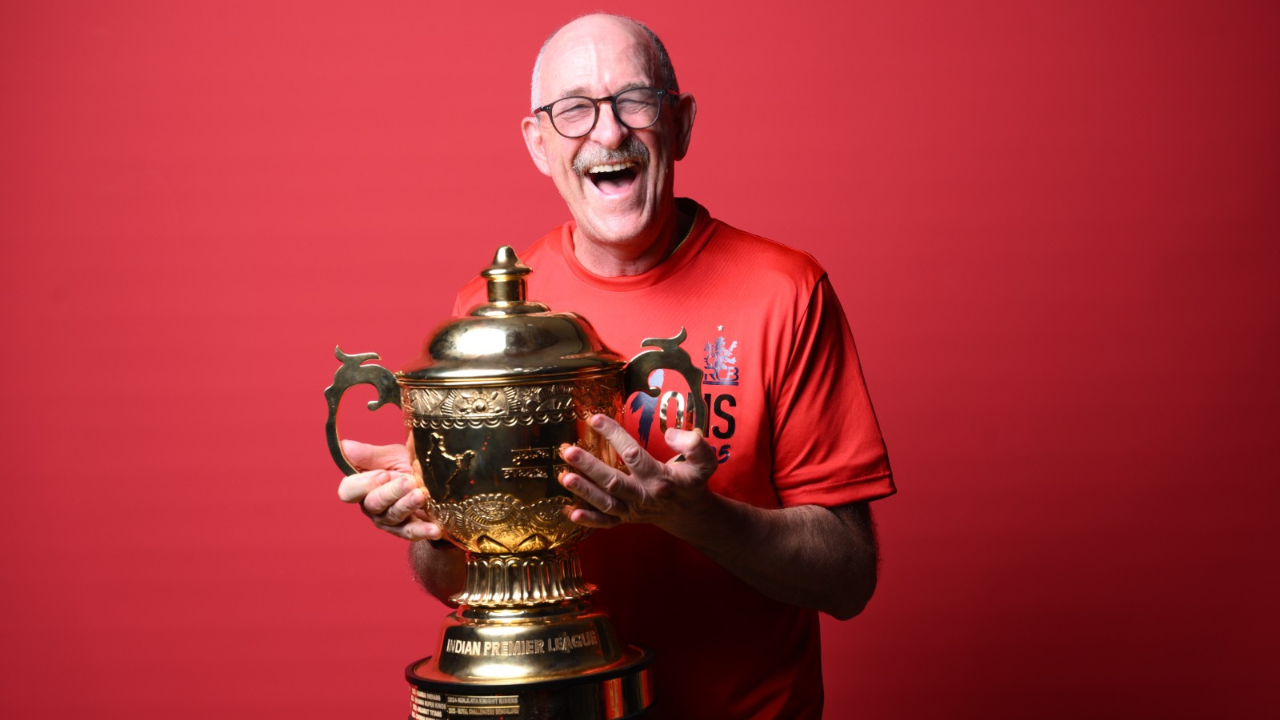 Evan Speechly, RCB physio, smiling while holding the IPL trophy against a red background.
