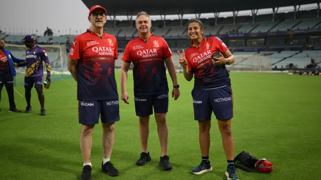 Evan Speechly with male and female RCB support staff standing on the cricket ground during an IPL training session at the stadium.