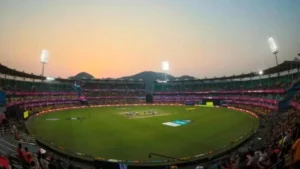 Wide view of a packed IPL cricket stadium during an evening match with floodlights on, spectators in the stands, and players on the field at sunset.