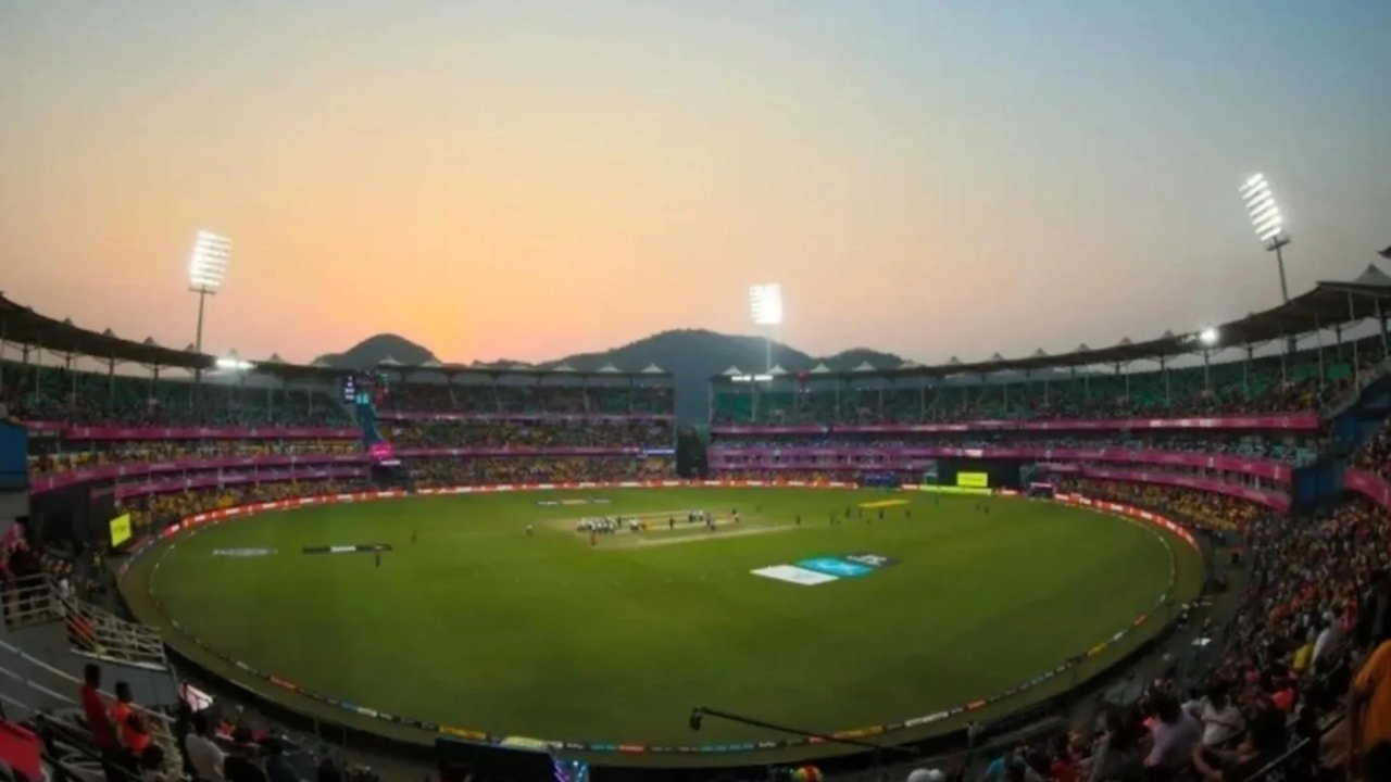 Wide view of a packed IPL cricket stadium during an evening match with floodlights on, spectators in the stands, and players on the field at sunset.
