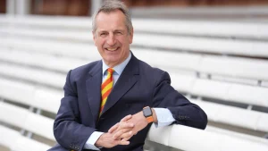 Portrait of Julian Metherell, the next President of MCC, seated at a cricket stadium wearing a navy suit and striped tie.