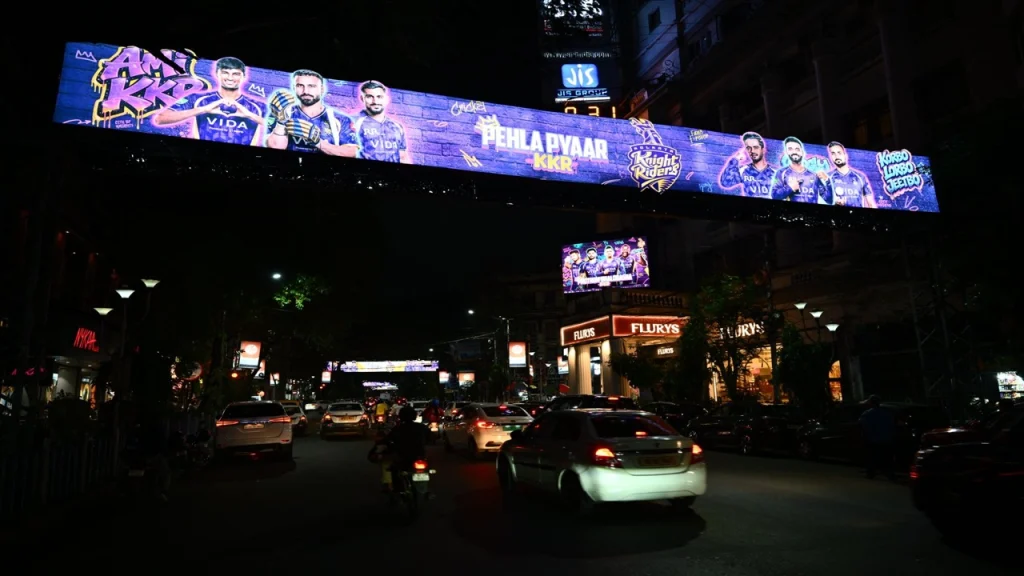 Kolkata Knight Riders IPL banner displayed over a busy Kolkata street at night with traffic and illuminated KKR players campaign artwork