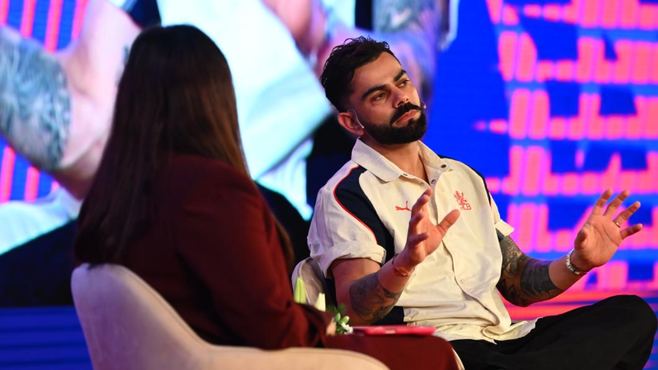 Virat Kohli speaking during an interview session at a public event while seated on stage with an interviewer and illuminated backdrop display.
