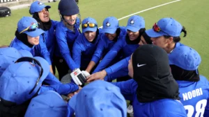 Women’s cricket team in blue uniforms gathered in a pre-match huddle on the field showing teamwork and unity before the game.