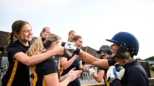 Women’s cricket players celebrating during a match with a batter wearing a helmet and teammates cheering after a successful moment.