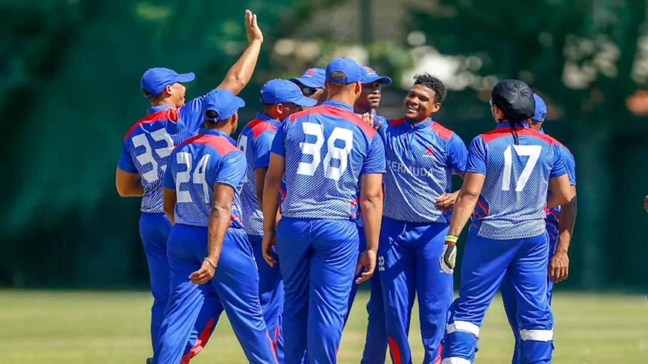 Bermuda national cricket team players celebrate a wicket during a cricket match, gathered together on the field in blue uniforms.