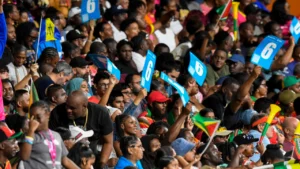 Fans in the stands cheer during a Caribbean Premier League match, holding six-run placards and waving national flags in a packed stadium atmosphere.