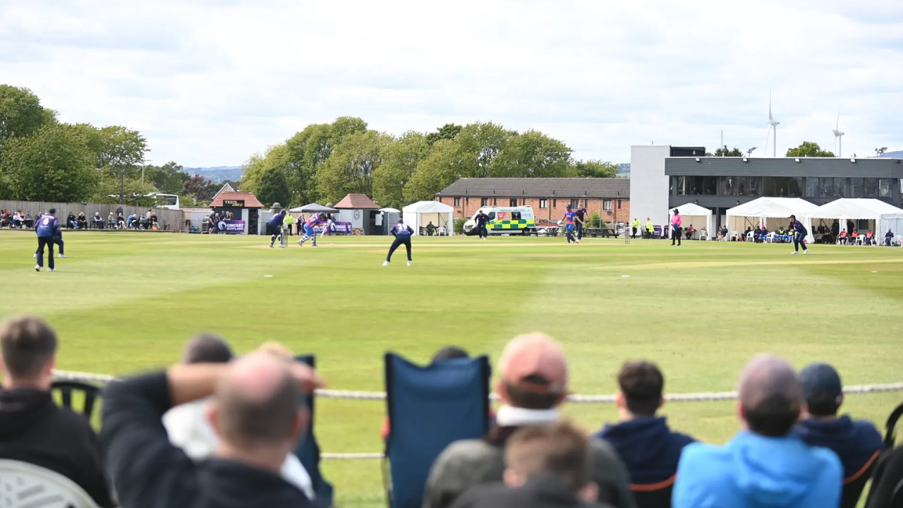 Spectators watch a live cricket match at a community cricket ground as players compete on a sunny day with fielders positioned across the outfield.