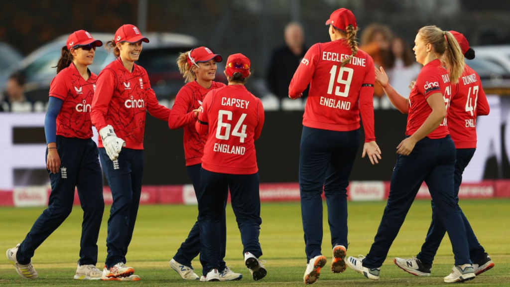 England women’s cricket team players celebrate a wicket during an international cricket match, with teammates gathering on the field in England’s red limited-overs kit.