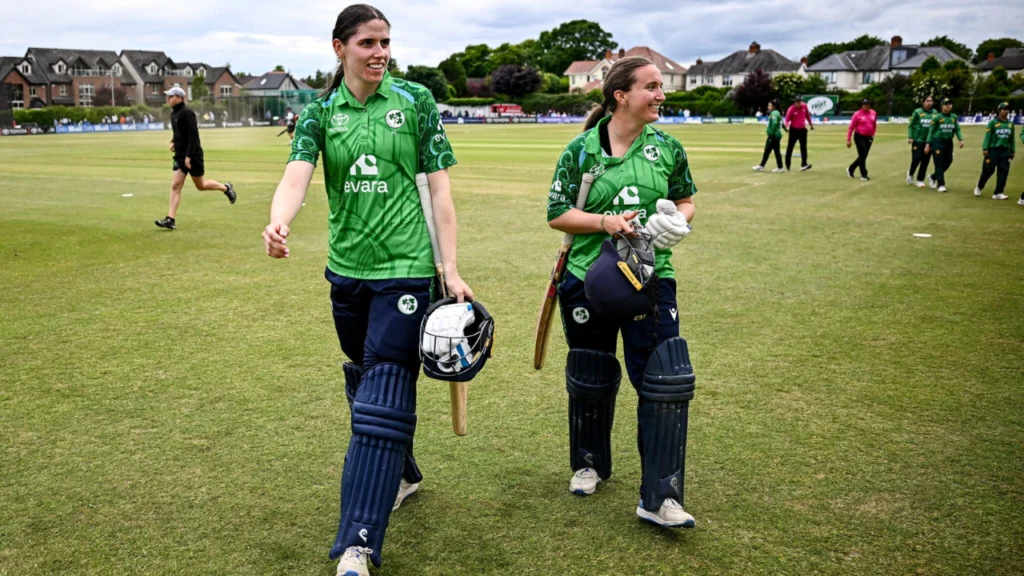 Two Ireland Women cricketers walk off the field with bats and helmets after a successful innings, celebrating a key batting partnership during an international match.