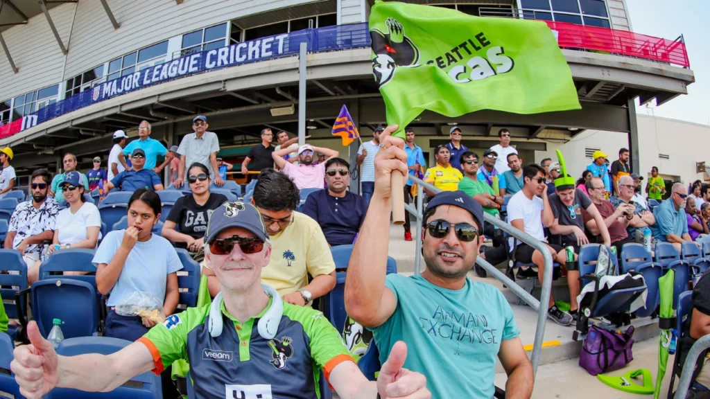 Seattle Orcas supporters cheer from the stands during a Major League Cricket match, waving team flags and showing support at a packed stadium.