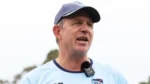 Shawn Bradstreet speaks during a cricket training session, wearing team apparel and a microphone while addressing players and staff outdoors.