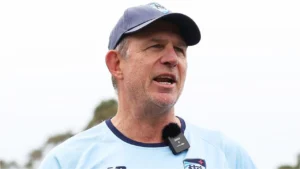 Shawn Bradstreet speaks during a cricket training session, wearing team apparel and a microphone while addressing players and staff outdoors.