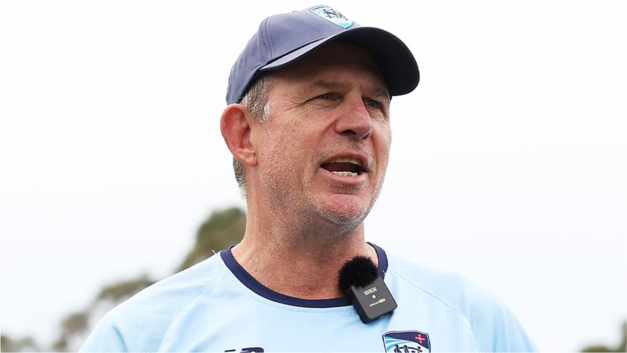 Shawn Bradstreet speaks during a cricket training session, wearing team apparel and a microphone while addressing players and staff outdoors.