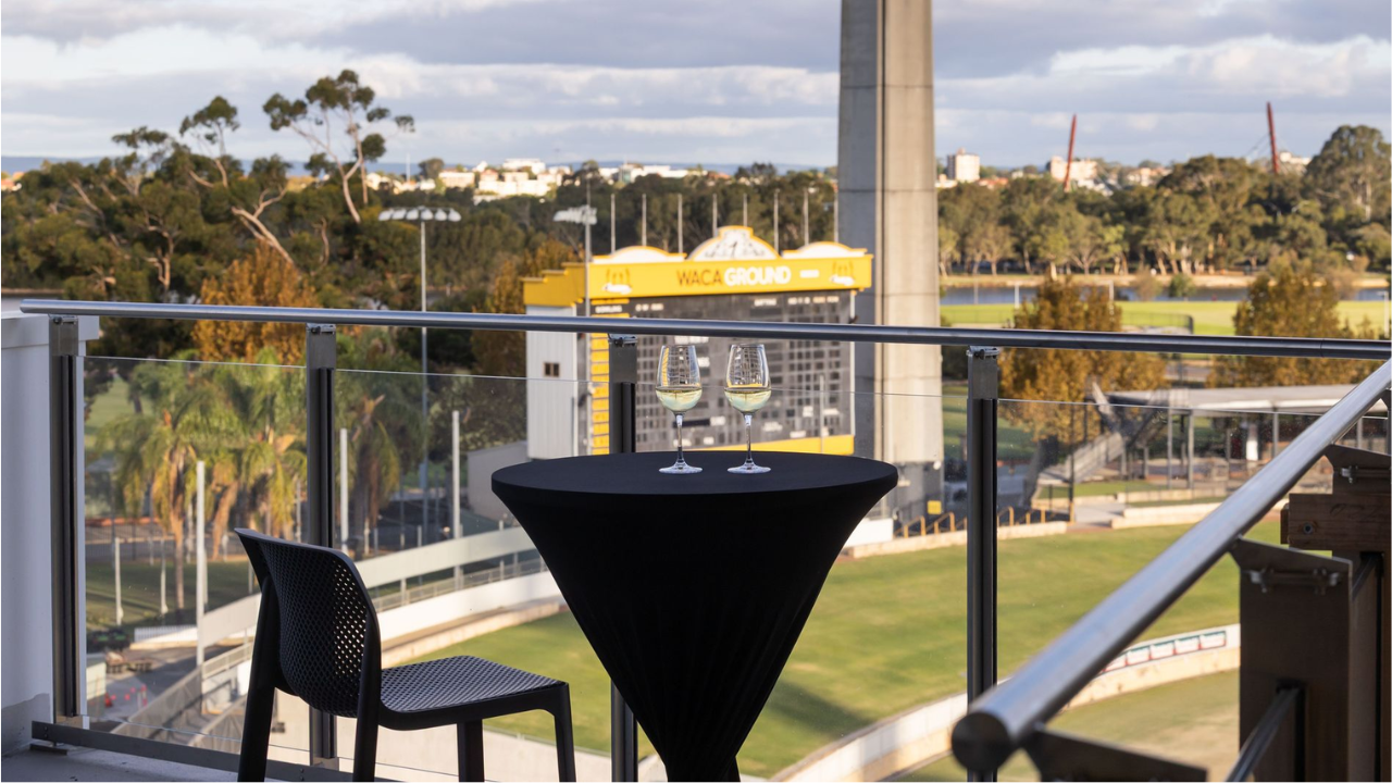 View of WACA Ground in Perth from a hospitality balcony with a table and two wine glasses overlooking the cricket stadium.