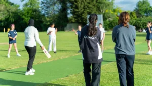 Coaches supervise a girls' cricket coaching session as young players practice batting and fielding on an outdoor cricket pitch.