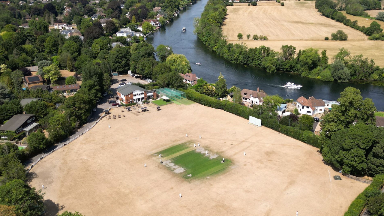 Aerial view of the scorched outfield at Maidenhead and Bray Cricket Club during dry weather, with the cricket pitch remaining green beside the River Thames.
