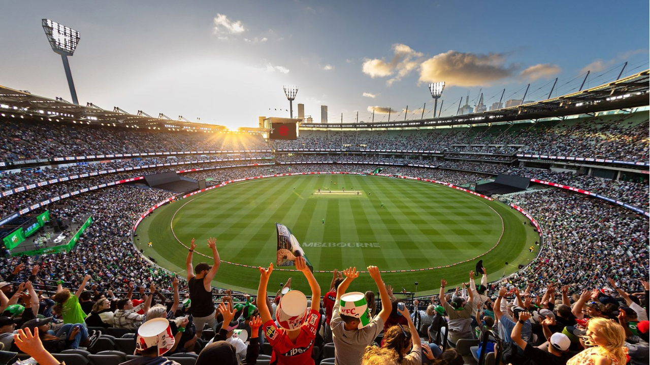 A packed crowd fills the Melbourne Cricket Ground (MCG) during a cricket match, with a panoramic view of the stadium at sunset.