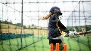 Young woman cricketer batting during a training session in outdoor cricket practice nets, wearing a helmet and protective gear while developing batting skills.