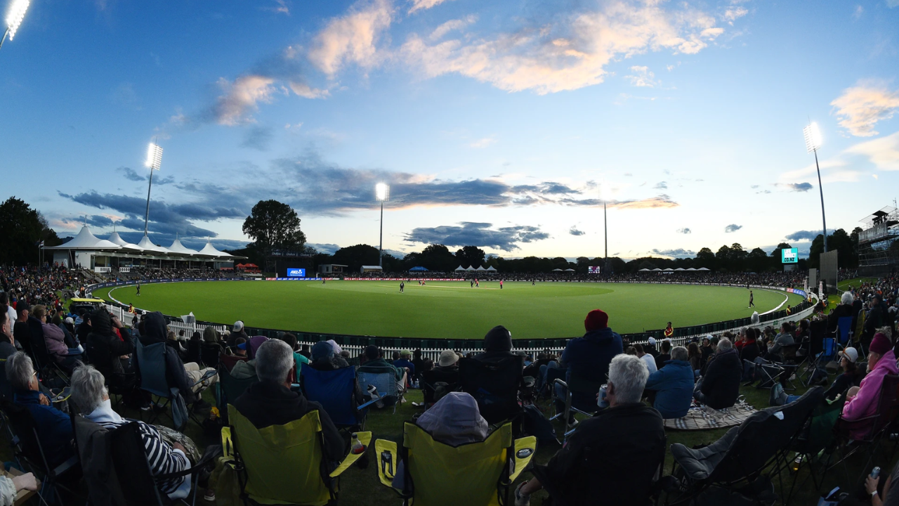 Crowd watches a cricket match under floodlights at a cricket ground in New Zealand.
