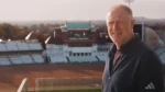 Alex O'Dowd smiling while standing in front of a cricket stadium, with the pavilion and seating visible in the background.