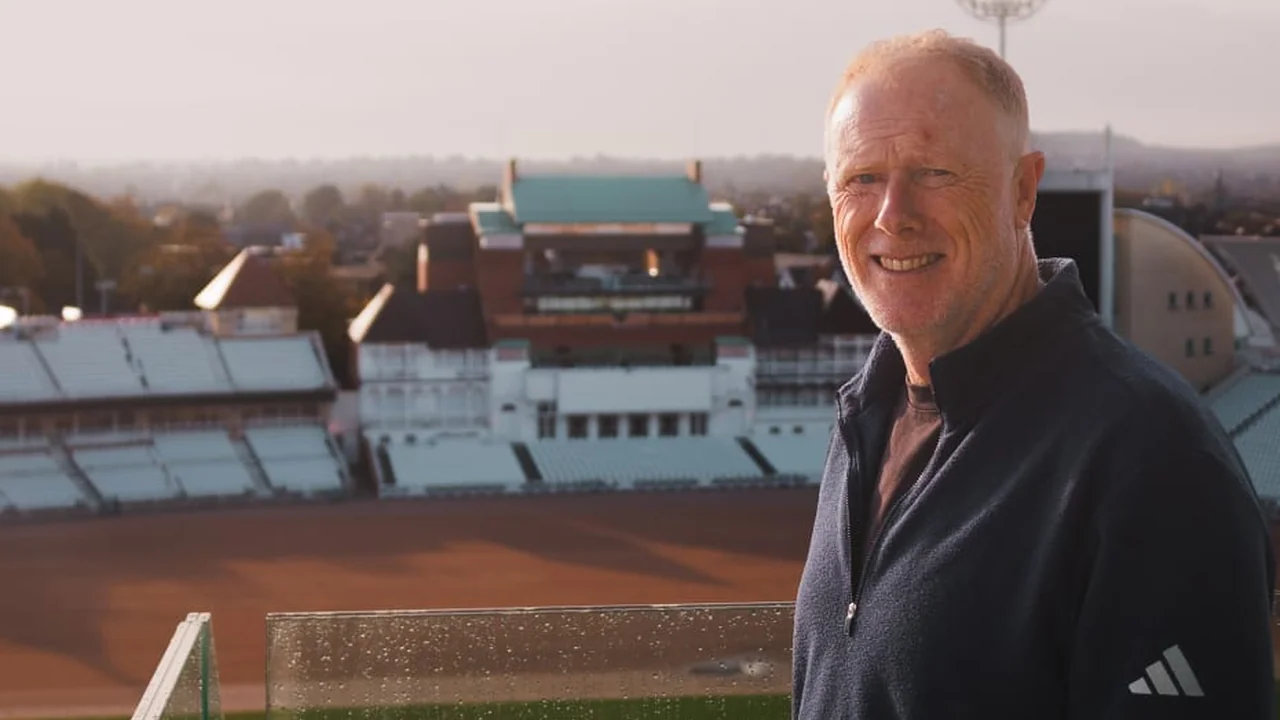 Alex O'Dowd smiling while standing in front of a cricket stadium, with the pavilion and seating visible in the background.