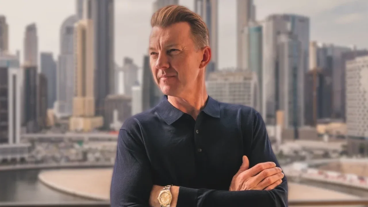 Former Australian fast bowler Brett Lee poses with his arms crossed, wearing a navy blue polo shirt against a modern city skyline.