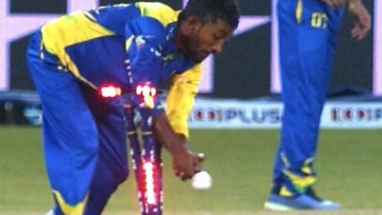 Cricket player in a blue and yellow uniform removes the LED bails from the stumps while holding a cricket ball during a match.