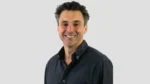 Portrait of Ed Cowan, former Australian Test cricketer associated with Cricket NSW, smiling in a dark blue shirt against a light grey background.