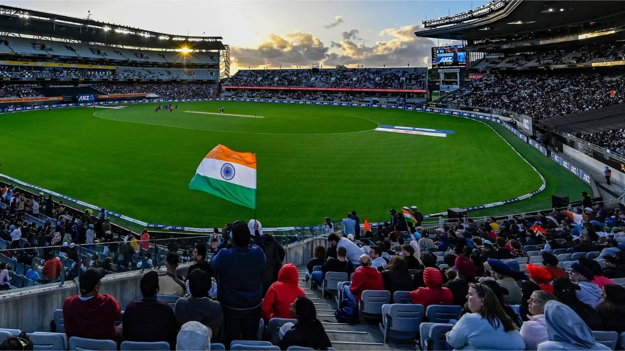 Cricket fans wave the Indian national flag in a packed New Zealand cricket stadium as spectators watch a match under the evening sky.