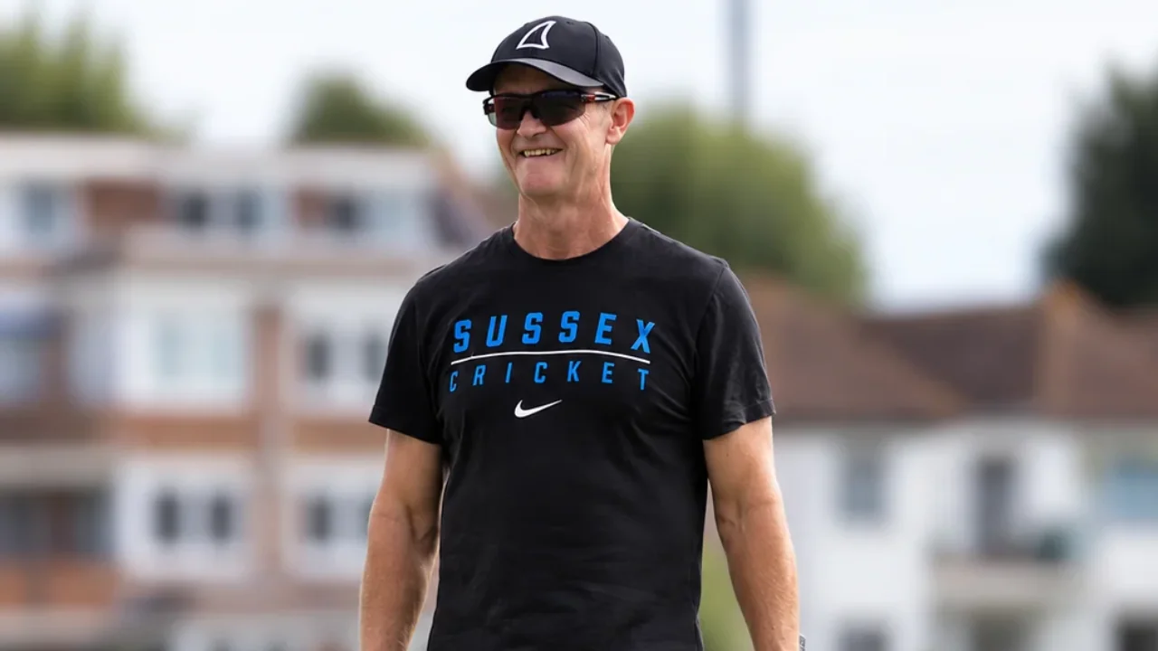 Grant Flower, Surrey County Cricket Club coach, walks across a cricket ground wearing Sussex Cricket training gear and carrying a cricket bat and fielding glove.