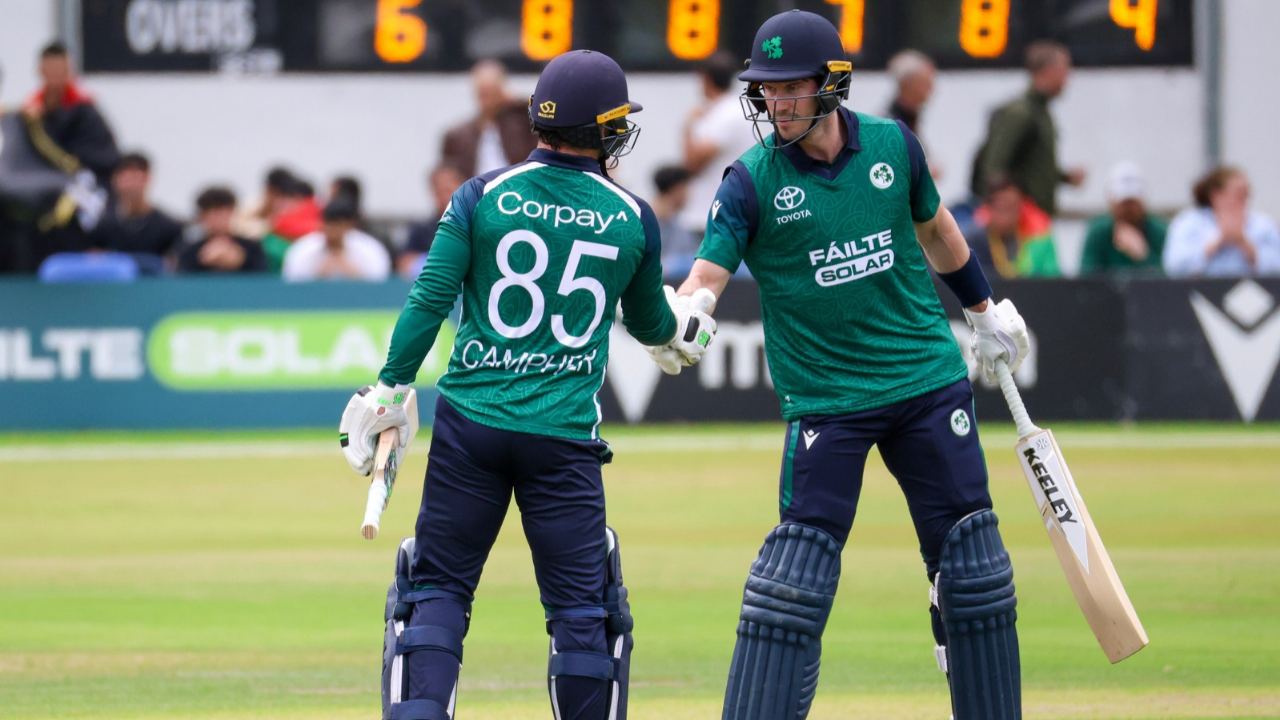Ireland men's cricket team batters celebrate a batting partnership with a fist bump during an international cricket match, wearing green jerseys.