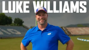 Luke Williams, cricket coach, smiles while wearing a blue Mumbai Indians polo shirt and cap at a cricket stadium.